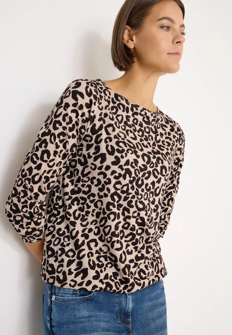Frau mit Leopardenmuster-Bluse und blauer Jeans steht an einer weißen Wand.
